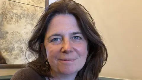 A woman with shoulder-length dark hair looking into the camera. She is wearing a dark jumper and has long gold earrings in. Behind her is a beige wall and a mirror
