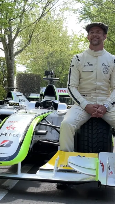 Jensen Button wearing wite racing overalls, sitting on the front wheel of a racing car.