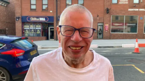 Jade Taylorson/BBC Stephen Tiller, in his late 50s, with very short grey hair and glasses and wearing a white t-shirt, smiles into the camera on a busy high street. 