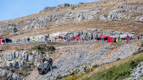 FPNW Upturned car on the side of a steep slope as emergency crews and vehicle recovery specialists work at the scene