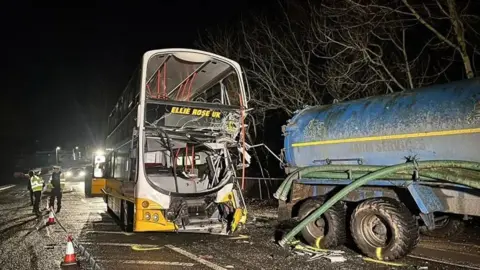 The double decker bus with both its front windows smashed and its back emergency door opened stands next to the end of blue tanker with surrounded by police tape and traffic cones with police officers in high visibility jackets standing by it