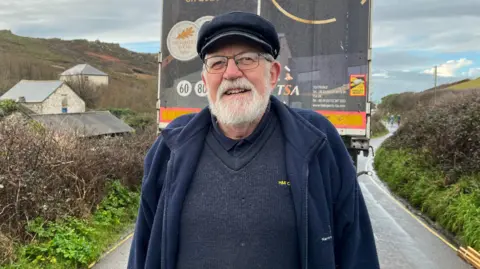 Barry Mundy is standing in front of the rear of the lorry. He is wearing a navy fishing cap and a navy fleece over a thick knitted navy jumper and navy shirt. He is smiling. He has a white beard and is wearing a pair of metal rimmed glasses.