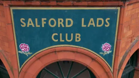 The green and gold-bordered Salford Lads Club sign, which has two red roses painted in the corners