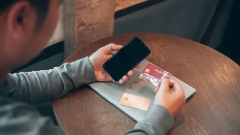 Getty Images Man holds a credit card and a phone with another bank card and a laptop on the table in front of him.