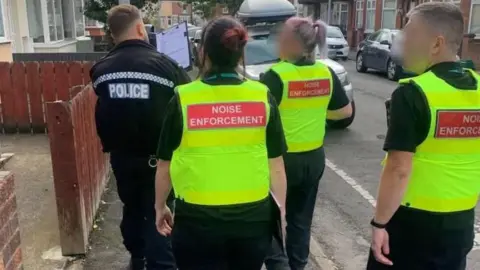 The backs of two female noise enforcement officers and one male one. They are wearing fluorescent yellow vests. A male police officer is on the left. They are walking along a residential street.