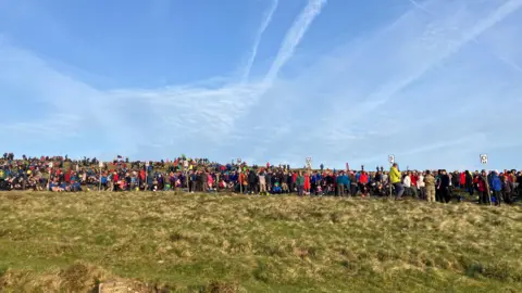 General view of participants setting off at Ten Tors in 2025. Hundreds of participants are stood in a long line on a grass field.