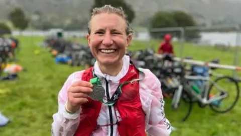 A woman with light brown hair standing in the rain in a green field, with bikes in the background. She is smiling and holding a medal around her neck. She is wearing a white waterproof jacket and red running vest, with a bib numbered 102 attached to her leggings.