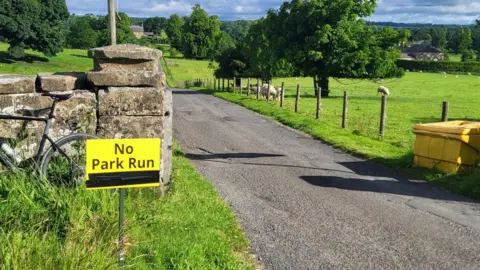 Kirkharle Courtyard Parkrun A luminous yellow sign reading "No Park Run" has been stuck into the ground next to the track of the park, which stretches through a grassy meadow with sheep grazing behind a wire fence.