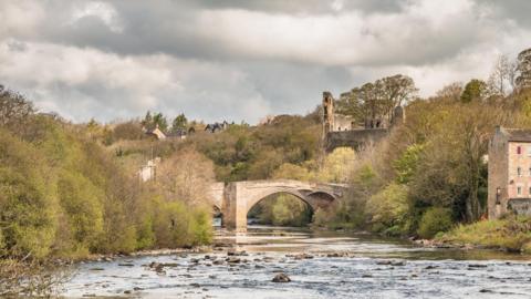 Photograph of a river with historic buildings of Bernard Castle in the background.  Cloudy, grey skies overhead.