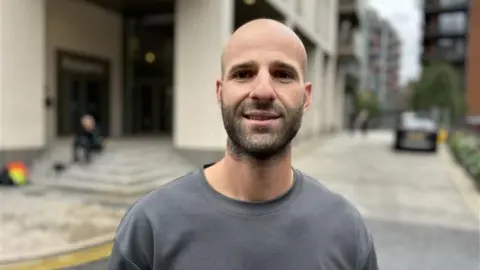 Yunus Mulla/BBC Nikolas Savva, who has a bald head and dark stubble, smiles at the camera wearing a grey jumper. 