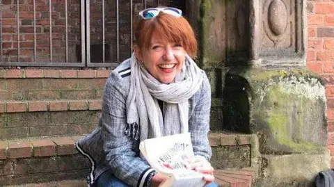 A lady with auburn hair is sitting on on stone steps and is holding a book that is half open. She is smiling into the camera and is wearing a grey scarf and jacket. 