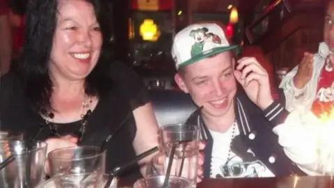 Roman Dibden A middle-aged woman with black hair smiles clutching a glass of water while sitting at a restaurant next to a young man in a cap who is also smiling.