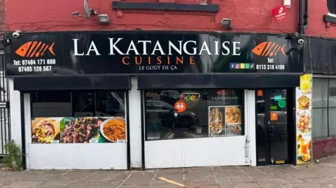 Daniel Pokas Street-taken shot of La Katangaise restaurant in Leeds. The restaurant is on a corner and has dark billboards above with the name of the restaurant's name spelt out in orange lettering. Pictures of the food is plastered on the windows.