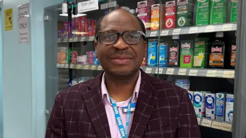 A head and shoulders shot of a man standing behind a pharmacy counter, smiling at the camera. He has short black hair on the sides of his head, and black stubble on his face. He is wearing tortoise-shell glasses, a pink shirt, a burgundy, checked blazer, and a blue NHS lanyard. Medications line shelves behind him.