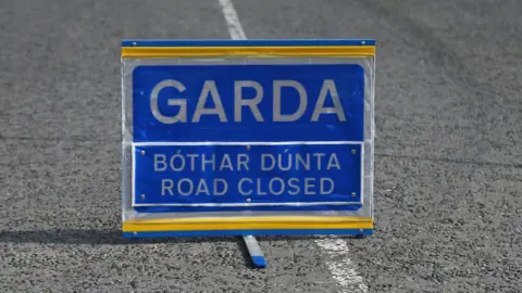 A blue sign placed on a road with white markings. The sign says Garda and road closed in both Irish and English. 