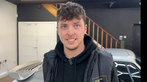 Jamie Morris A man with dark brown curly hair and small beard facing the camera. He is wearing a black top and and a puffa jacket. There is a car behind him and a flight of stairs.
