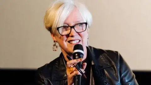 Janice Forsyth in a leather jacket and wearing dark plastic glasses speaking at an event. She is holding a microphone and smiling, her hand waving in the air.