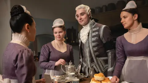 Netflix Three women wearing purple maid outfits, all have brown hair wrapped in a bun with a small hat on top. There is also a man wearing grey footman attire and has blonde hair. They are standing around a table that has grey silverware and scones on it.