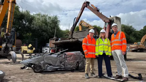 Wiltshire Council Three people in high-vis jackets and white helmets are pictured standing in front of a crushed grey-coloured car and what looks like some kind of mechanical crushing device. There is also a digger with a claw to the extreme left of the image. It is a cloudy day. The people pictured all look relatively serious.