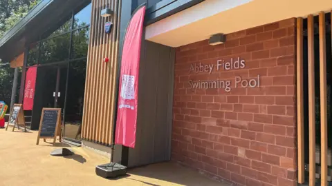 The outside of a brick building with glass doors and wooden cladding on parts. On the red brick wall are the words Abbey fields Swimming Pool in silver letterin.
Red flags fly outside which say Warwick District Council and Everyone Active.