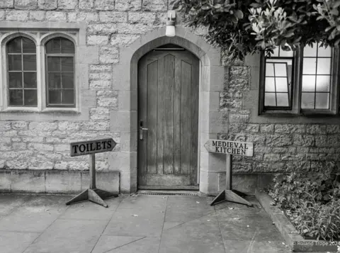 Roland Trope Signs reading "Toilets" and Medieval Kitchen" point to a door in a wall