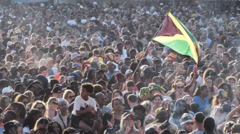 A huge crowd of people at a festival. A Jamaican flag is flown prominently.