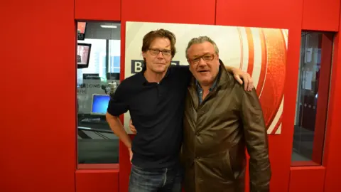 Robert Elms wearing a black polo shirt standing next to Ray Winston who is wearing a leather jacket