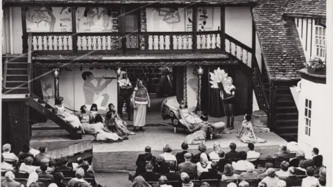 Shakespeare at the Towers A black and white photograph showing actors on an outdoor stage performing to an audience at the George Hotel. 