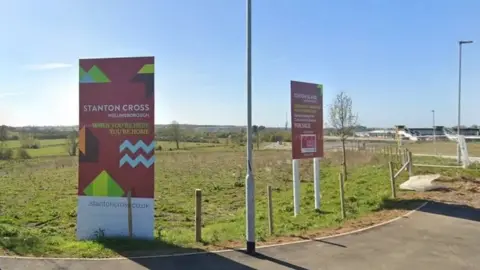 Two large red signs in the ground saying "stanton cross". They are in the grass with fields behind them.