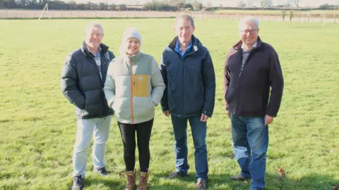 Malmesbury Town Council Three men and one woman stood smiling widely on a grass field. They are wearing winter clothes, with big coats and fleeces on.