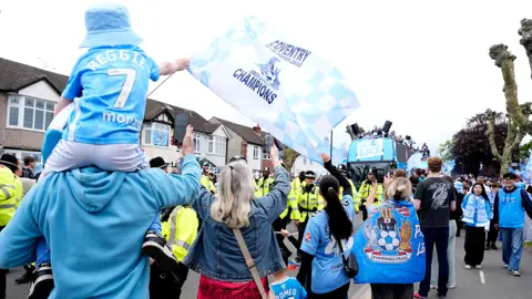 PA Media Fans lining the street as the bus approaches, with police in front of it in high-vis jackets. One young fan in the foreground on the left is on an adult's shoulders