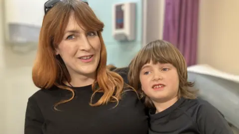 Danielle with her son Leo. Danielle has long red hair and Leo has short brunette hair. They are both wearing black. Leo is leaning against his mother's side and Danielle is looking at the camera. Both of them are smiling.