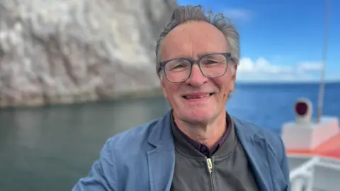 Sir Hew Dalrymple, wearing a blue jacket and glasses, on a boat beside the Bass Rock.
