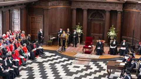 University of Cambridge Rows of college officials, dressed in red robes, look on as Lord Smith stands and speaks at the front. There are people seated to his side. The formal room is panelled in wood.
