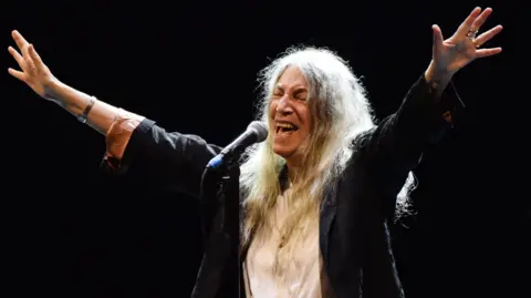 Patti Smith with her arms outstretched and singing on stage with a black backdrop. Dressed in dark jacket with white shirt. 