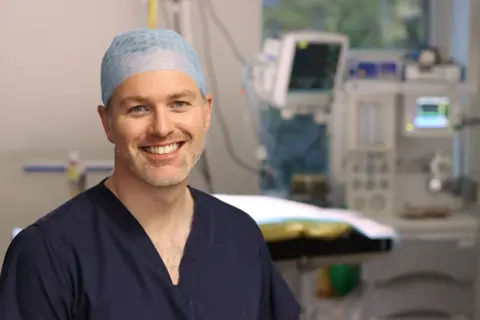 Mark Morton A man in a surgery room smiles, wearing a hair net and navy medical scrubs.