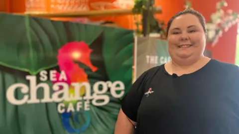 Munaza Rafiq/BBC Lexie is smiling directly into the camera. 
Her hair is pulled back into a ponytail and she is wearing a black  short sleeved t-shirt with the Sea Change logo. She is stood in front of a colourful board that says Sea Change cafe. The logo is a mulit-coloured seahorse.