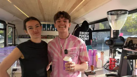 Luke Derbyshire coffee shop owner in pink T-shirt with female colleague in the coffee bus kitchen area near coffee machine smiling and laughing.