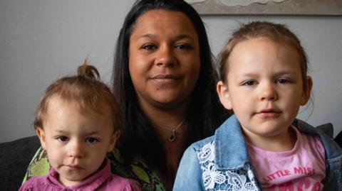 Chloe Bloomer, centre, a black woman in her thirties with long black hair looks straight at the camera, holding her two young daughters