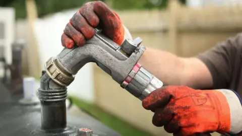 Gloved hands hold the nozzle of an oil hose over what looks like a filling hole of a domestic oil tank. Behind is a small strip of grass and an unpainted wooden fence.