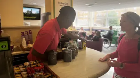 Northamptonshire Health Charity Volunteer in red top behind a cafe counter serves a woman wearing a pink shirt