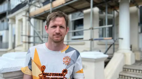 Builder Anthony Harman stands in the foreground looking at the camera. He has short light brown hair and a goatee beard. He wears a sports shirt that's white with grey and yellow patterns. Behind him is a house covered in scaffolding with steps approaching the front door. 