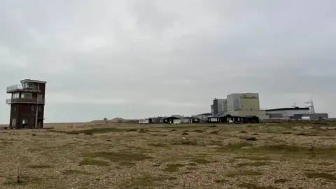 Coastguard lookout station on Dungeness beach with a nuclear power station in the background and a shingle beach in the foreground.