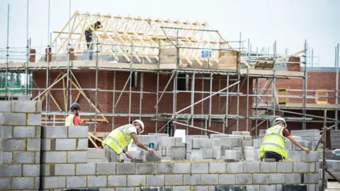 In the foreground, two men in yellow high-vis vests are laying concrete grey bricks. In the background is scaffolding around a brick house with the wood battens of the roof exposed.