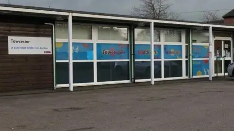 A one storey flat roof building with lots of windows at the front. A white sign that says Towcester Sure Start Children's Centre. There is a large car park in front of the building.