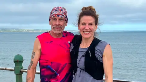 Two runners posing for a photo in front of the ocean, a man in a bandana and a woman in a runner's gilet