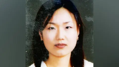 Jong-Ok Shin in what appears to be a copy of a family photo, where the folds and marks on the print are still visible, showing a close-up of a young women with long, dark hair and small hooped earrings looking straight at the camera with a neutral expression.