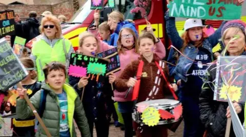Debbie Burrows Adults and children march along a street carrying colourful placards. A girl in the front of the photo is carrying a drum and drumsticks.