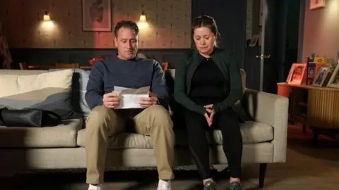Lime Pictures Diane Hutchinson, played by Alex Fletcher, and Tony Hutchinson, played by Nick Pickard, are sitting on a couch in a living room and appear emotional. Tony is holding a piece of paper in his hands with information of Diane's diagnosis.