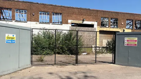 Tony Fisher/BBC Grey hoardings and a black gate in front of a derelict building which has vegetation growing outside it. Windows are smashed and an entrance is boarded up with plywood. There are signs on the grey hoardings which read "Danger - keep out".